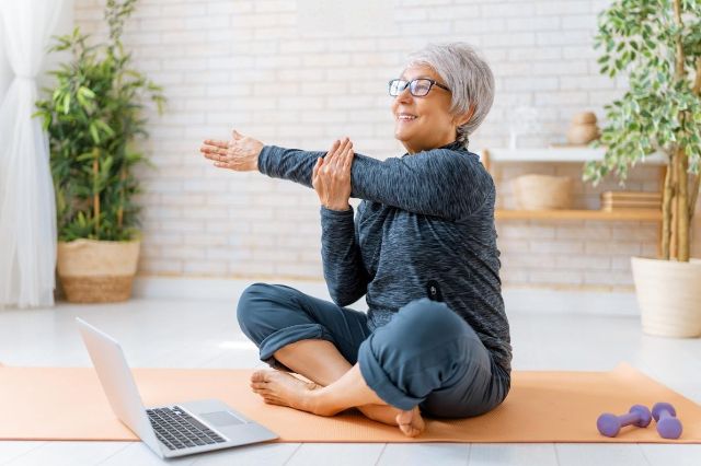 Seniorin sitzt auf Yogamatte vor ihrem Laptop und nimmt an einem Kursangebot online teil.
