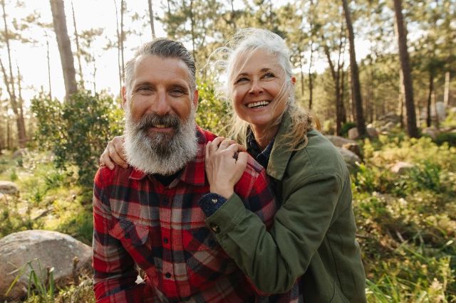 Ein älteres Paar befindet sich umarmend in einem Wald und lächelt in die Kamera.